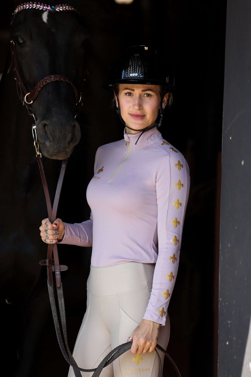 Woman wearing dusty purple performance riding top with gold fleur-de-lis designs, holding horse reins