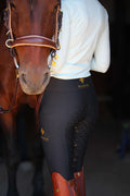 Close-up of a rider wearing black Masego Riding Tights with silicone grip and gold logo, standing next to a brown horse.